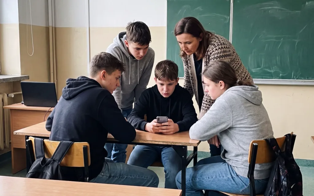 Czworo nastolatków i nauczycielka w klasie z zieloną tablicą pochylają się nad smartfonem, który trzyma chłopiec przy ławce